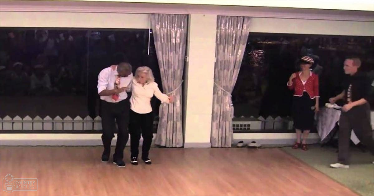 Dance scene with four adults indoors; two holding wands or sticks against large windows and curtains at dusk.