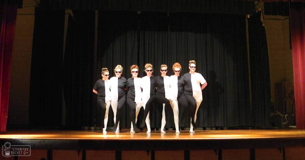 Young adult female performers wearing black tops, white pants or skirts, dark sunglasses, and white sneakers on stage in front of a dark curtain backdrop.