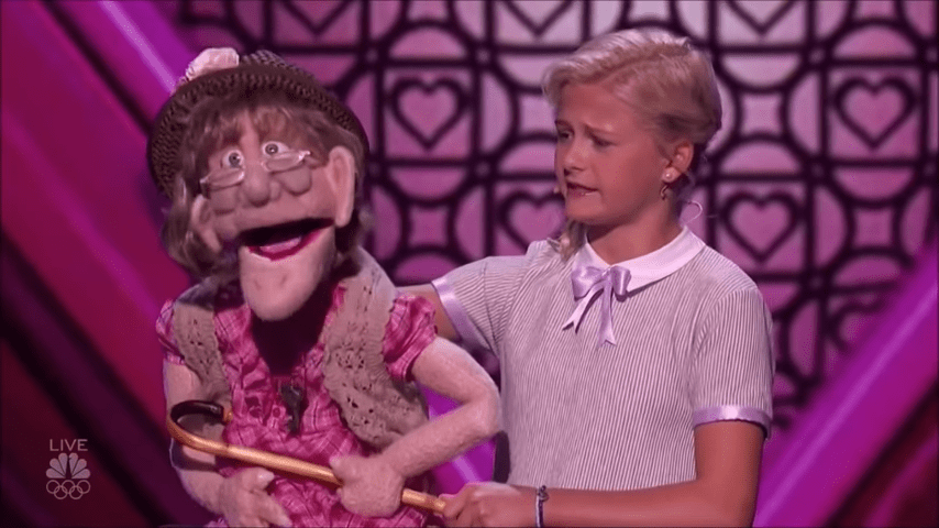A puppet character in a pink dress with ruffled sleeves, held by an adult female puppeteer against a backdrop of heart shapes and bright studio lighting.