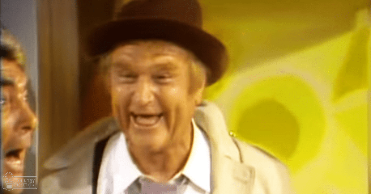 An older adult male wearing a light-colored jacket with a white shirt and dark hat stands indoors against a bright yellow background.
