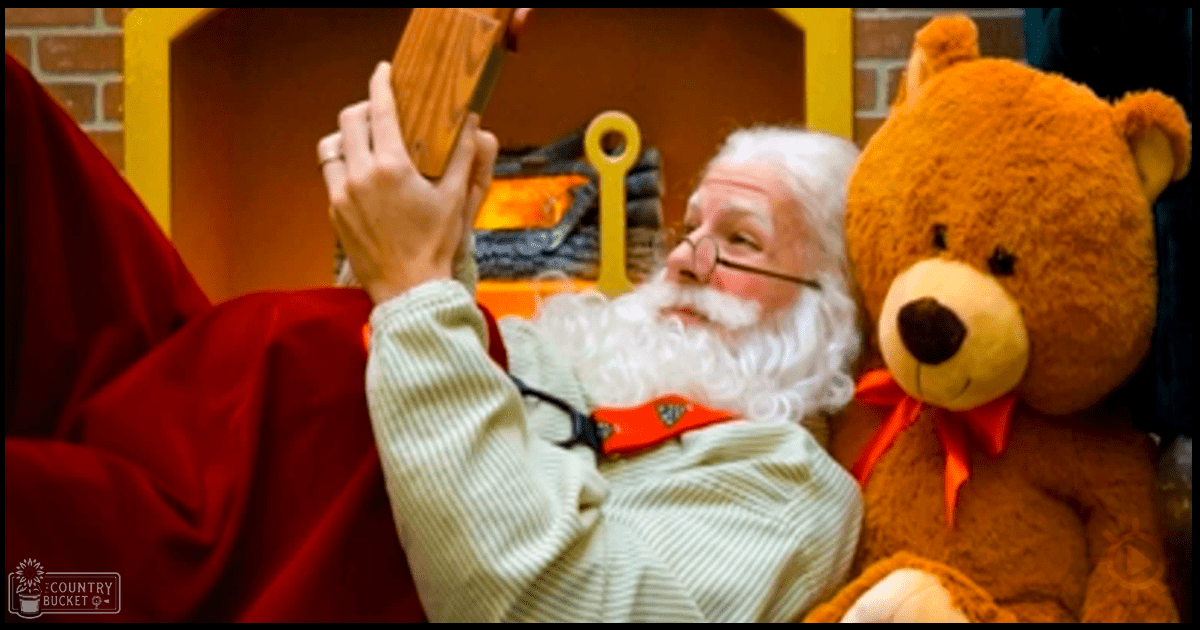 An older adult male dressed as Santa Claus in a cozy indoor setting, holding a wooden block with a stuffed brown teddy bear wearing an orange bow tie and a yellow toy shovel handle visible in the background.