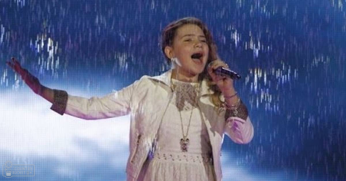 A young female child, approximately 8-10 years old, performs on stage wearing a light-colored dress with lace and buttons, holding a microphone against an artificial rainfall backdrop.