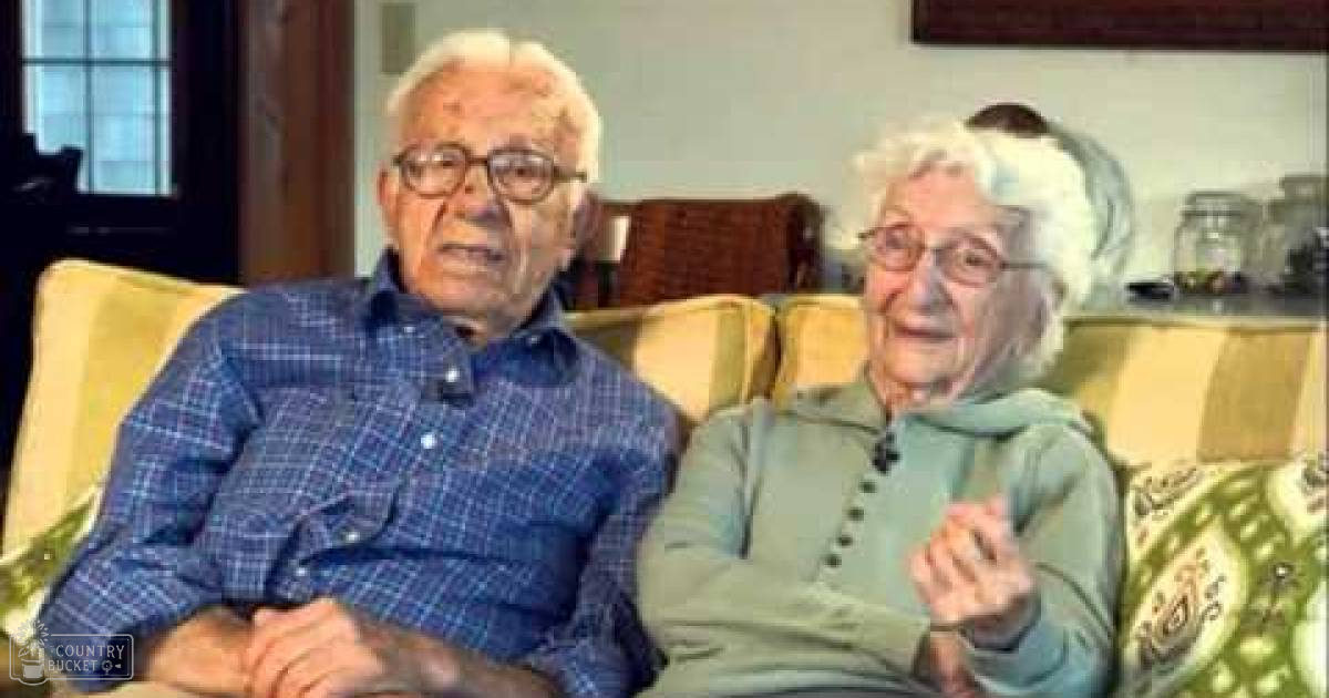 An older adult couple wearing glasses and casual clothing in a living room setting with beige furniture, natural light filtering through windows.