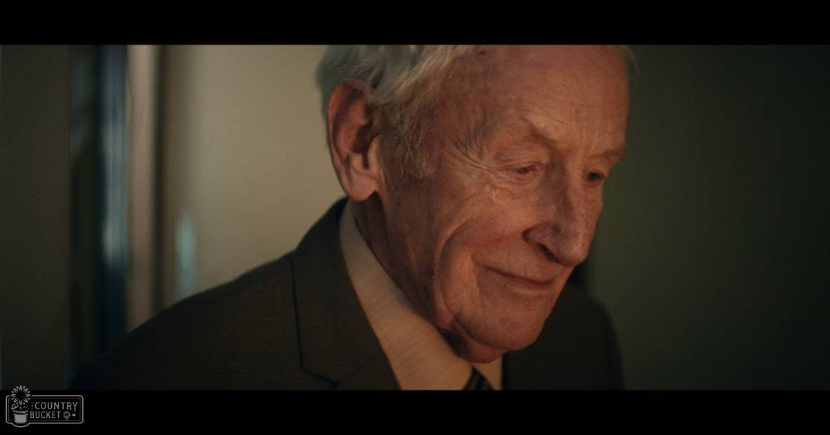 An older adult male wearing a dark-colored suit jacket and light collared shirt indoors during daytime, with neutral-toned walls as background.