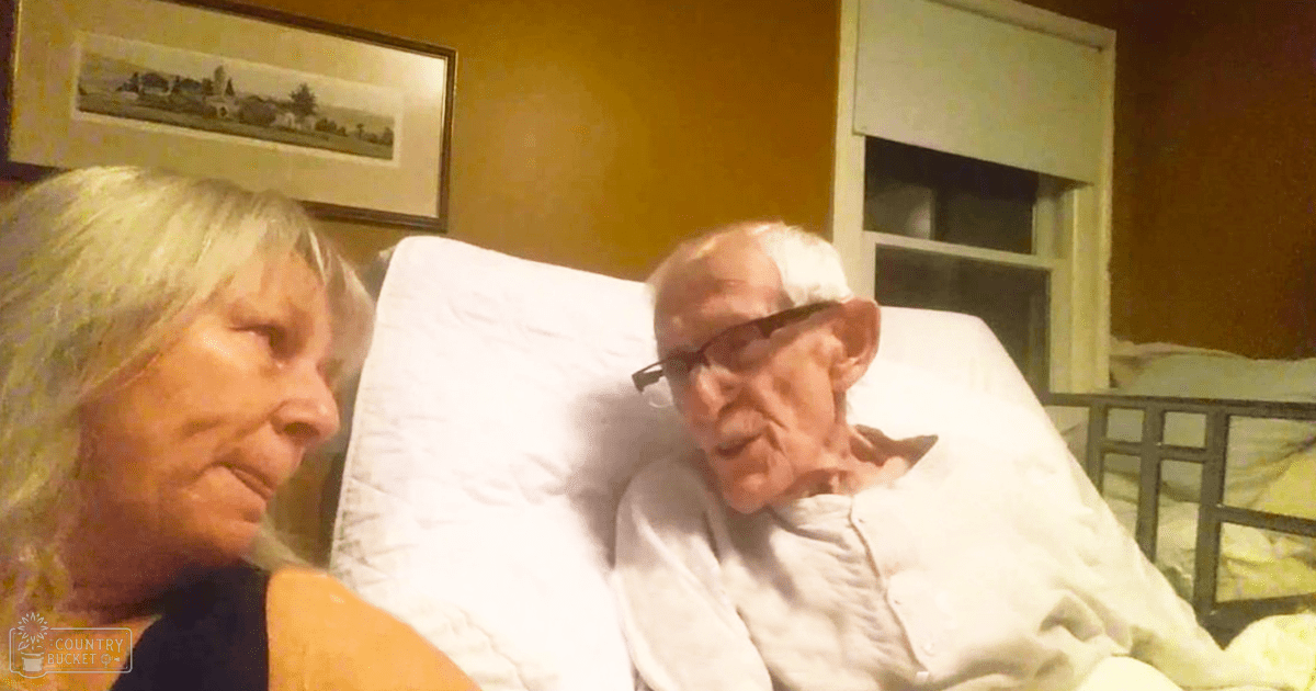 An older adult male in a hospital bed with an older adult female sitting beside him, indoors, evening or nighttime lighting