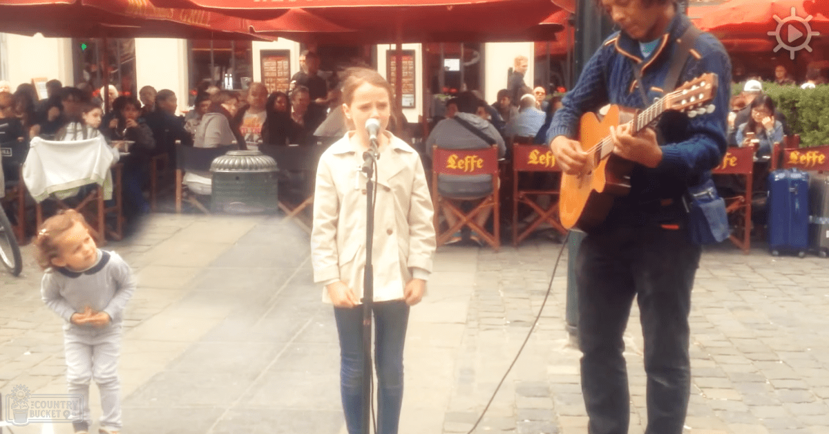 A young adult female singer performs outdoors in a café setting, accompanied by an adult male guitarist and an infant. The scene includes tables with red 'Leffe' branding and stone-tiled pavement.