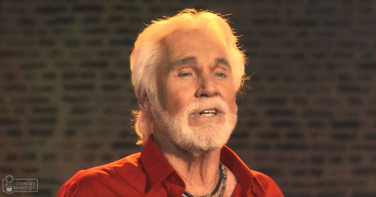 An older adult male wearing a red collared button-up shirt, indoors with controlled lighting and a patterned backdrop.
