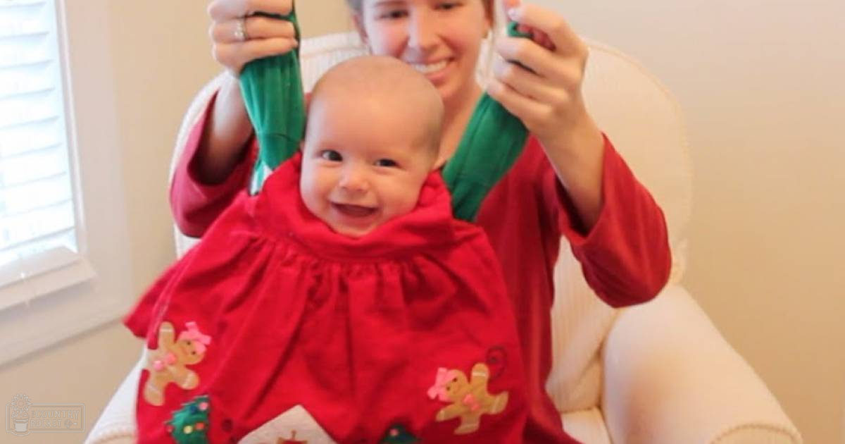 An infant wearing a festive red dress with gingerbread men and Christmas trees, held by an adult female in a home setting with natural light streaming through partially open blinds.