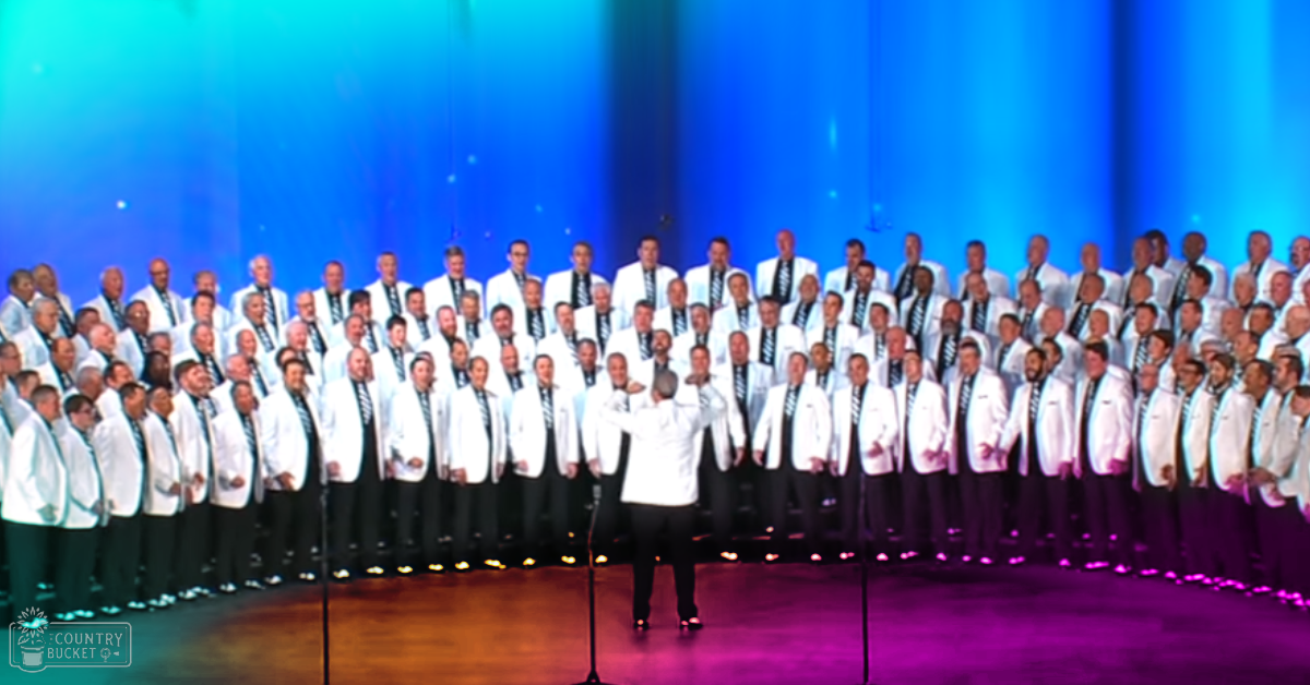 A choir of adult male performers in formal attire, white jackets with black ties, dark pants and dark shoes, performing on a stage with blue lighting effects in an indoor concert hall.