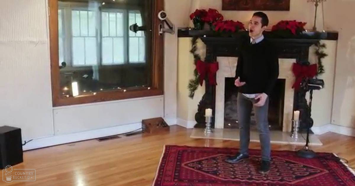 A young adult male in a cozy living room decorated for Christmas, wearing a black sweater over a light blue collared shirt with dark jeans and black shoes.
