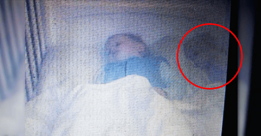 A young child in white pajamas lies in bed indoors with dim lighting and white bedding, suggesting evening or nighttime. A curtain is visible on the left side.