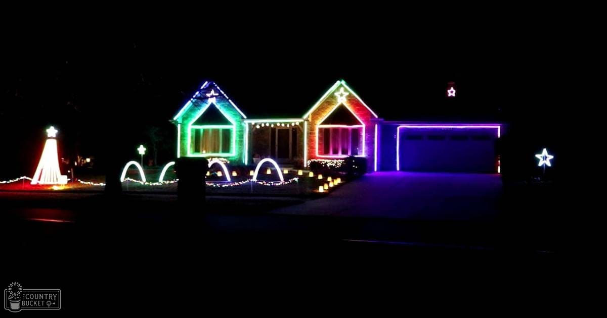 A residential neighborhood adorned with colorful holiday lights at night; a tall white light structure resembling a Christmas tree is visible.