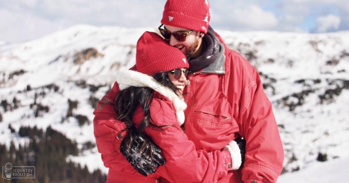 A couple in a snowy mountain setting; male adult wears red beanie, sunglasses with dark lenses, and red winter coat; female adult sports red knit hat, puffy jacket with fur-lined hood, black gloves.
