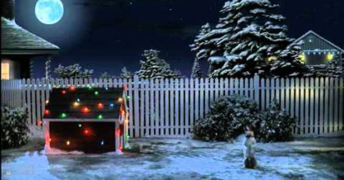 A snowy nighttime scene with a house, fence, snow-covered trees, decorated Christmas lights on a garage door, illuminated windows suggesting early evening or dusk in a residential neighborhood.