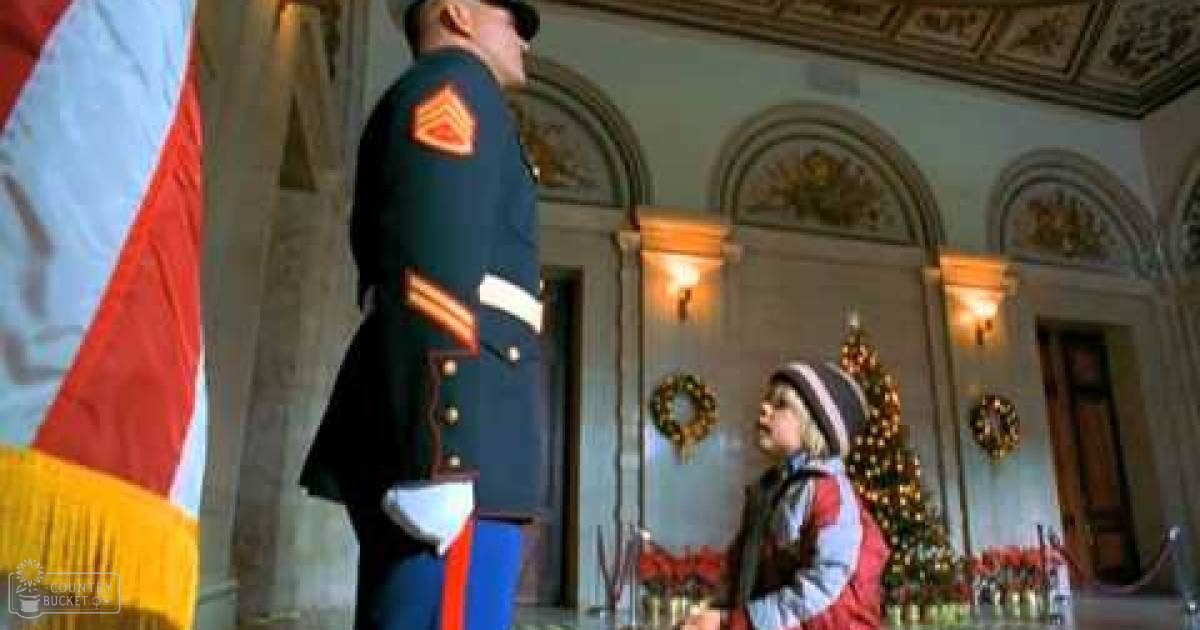 A toddler wearing a red-sleeved winter jacket and a knit hat, standing indoors with classical architecture and festive decorations around Christmas time.