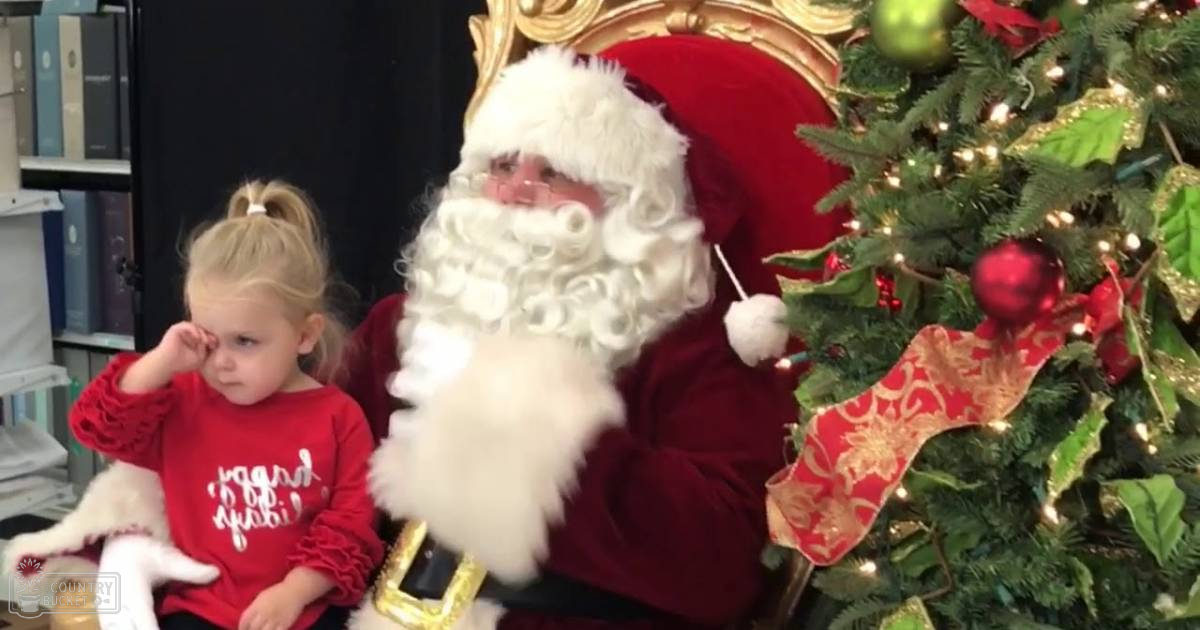 A child in a red 'Good Vibes' shirt interacts with Santa Claus inside, surrounded by Christmas decorations and a tree adorned with ornaments.
