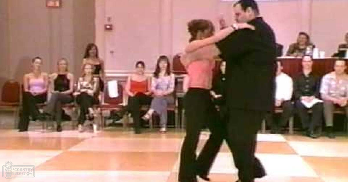 A couple dancing in an indoor dance hall or community center, surrounded by seated audience members watching the performance.