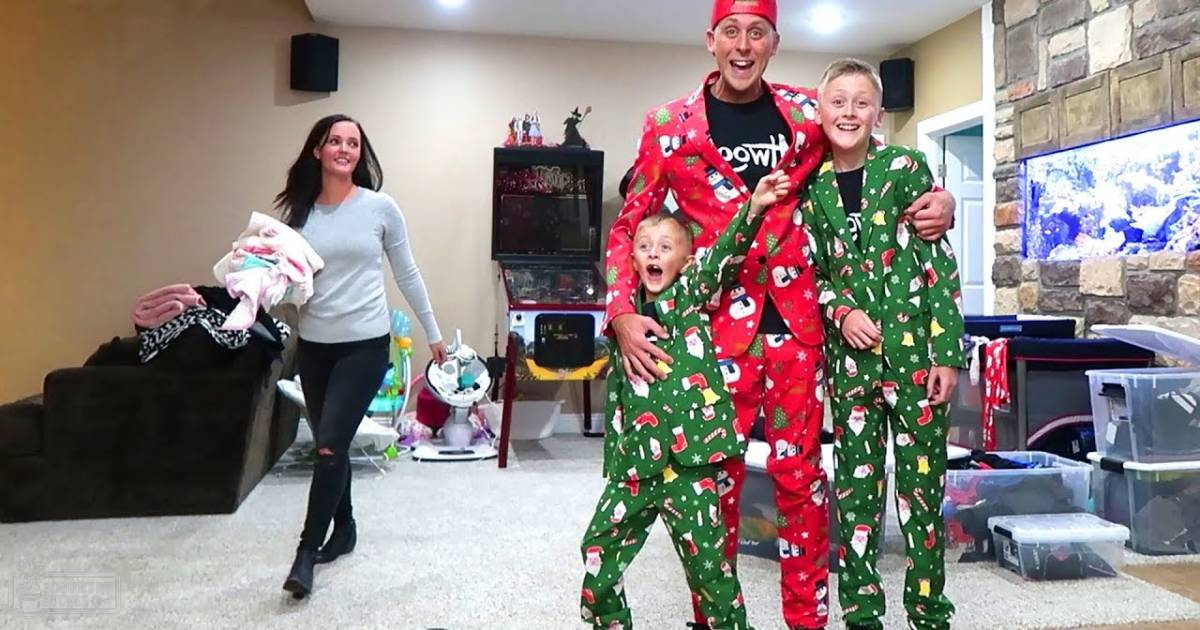 Adult Female carrying clothes, wearing light gray top and black pants with rips; Adult Male in red Santa suit jacket over t-shirt and green pajama-style pants; Child in green Santa-themed pajama set standing near adult male; Teen/Young Adult Male in similar attire to the adult male. Scene includes dark brown couch, arcade machine, television showing colorful display, plastic storage bins, baby swing with toys attached.