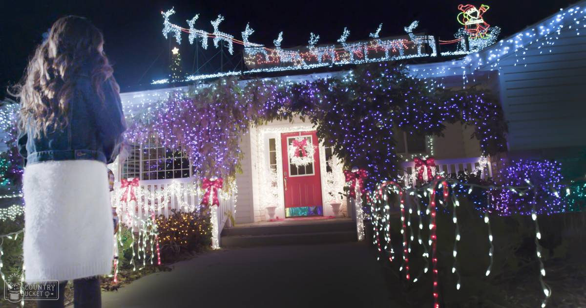 A young adult female in a denim jacket and white fluffy skirt stands in front of a house decorated for Christmas at night.