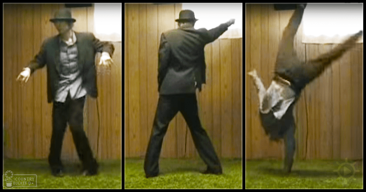An adult male dancer in a dark blazer with a light-colored patterned shirt, black pants, and fedora hat, performing a dance move outdoors on grassy ground with a wooden fence backdrop during soft late afternoon light.
