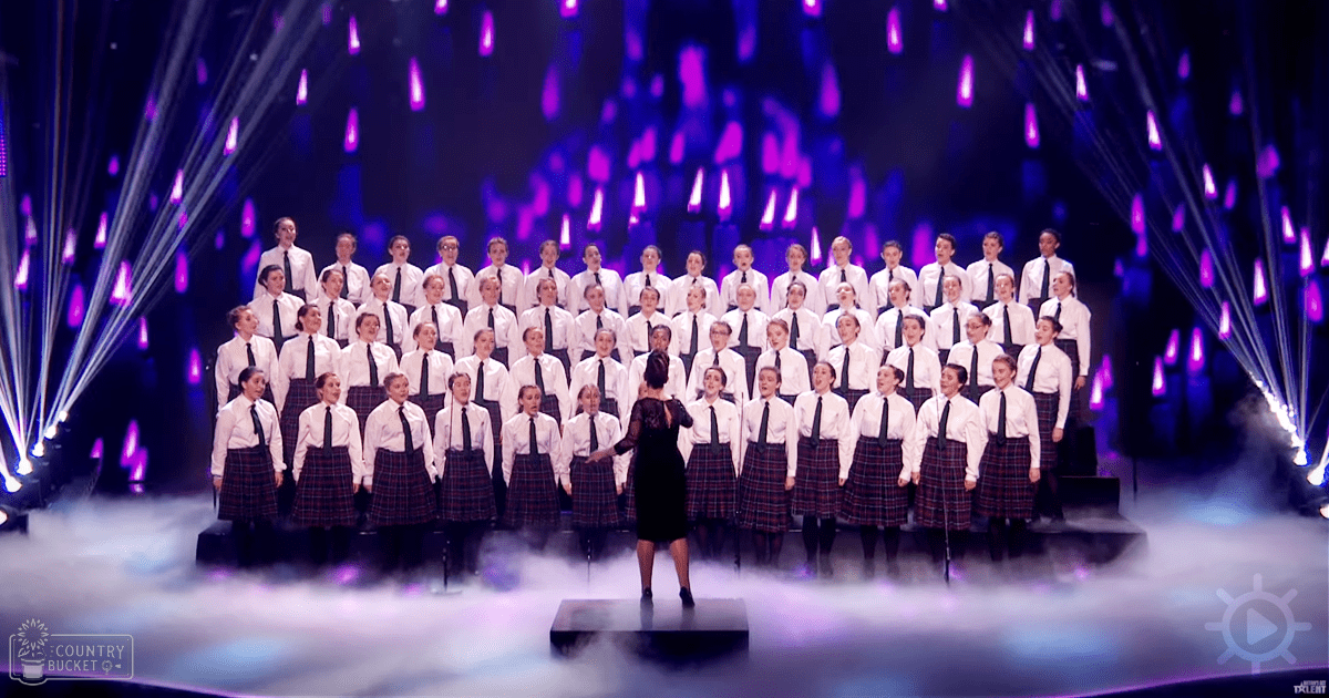 Teenage female choir performing on an indoor stage with purple lighting effects, emphasizing a school uniform in white shirts and plaid skirts.
