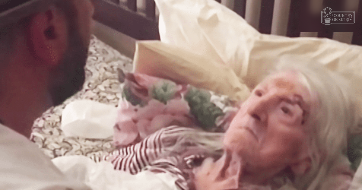 An elderly woman lies in a hospital bed indoors under natural light, wearing a pink and white striped garment with a low-cut neckline.