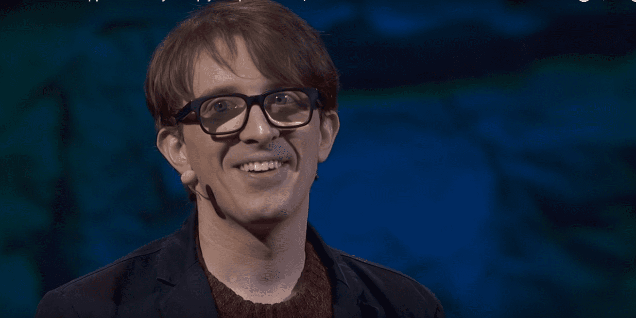 A young adult male in a relaxed fit dark blue or black collared shirt, wearing glasses with dark frames and a small microphone clipped to his collar, stands against a backdrop of abstract patterns on a dark blue screen, possibly during an indoor presentation.