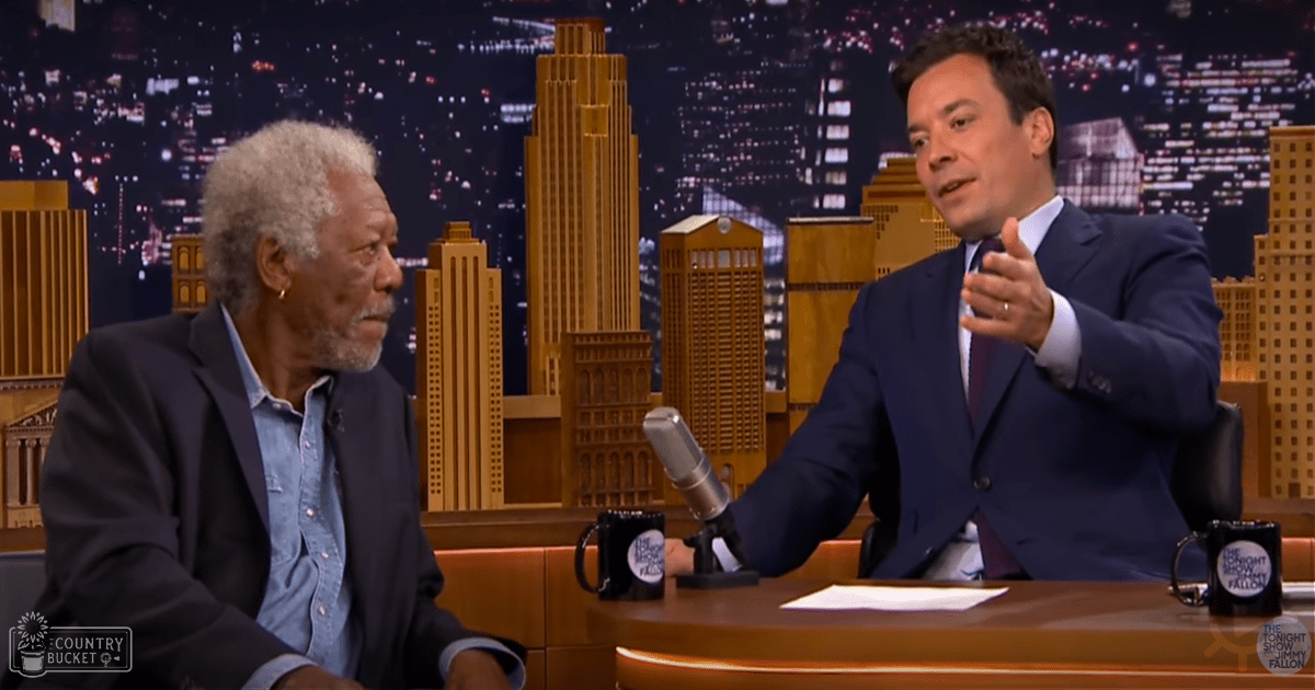 Talk show host in dark blue suit interviewing older adult guest wearing light denim shirt and dark gray blazer, set against a nighttime cityscape backdrop with studio lighting.