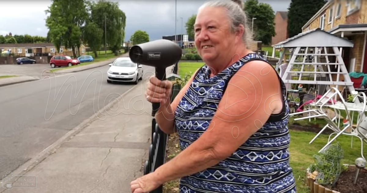 A middle-aged woman in a blue and white patterned sleeveless top holds a black hairdryer, standing outdoors by a white metal garden table with chairs in the background. A white lattice gazebo and parked cars are visible in an outdoor residential area on a sunny day.