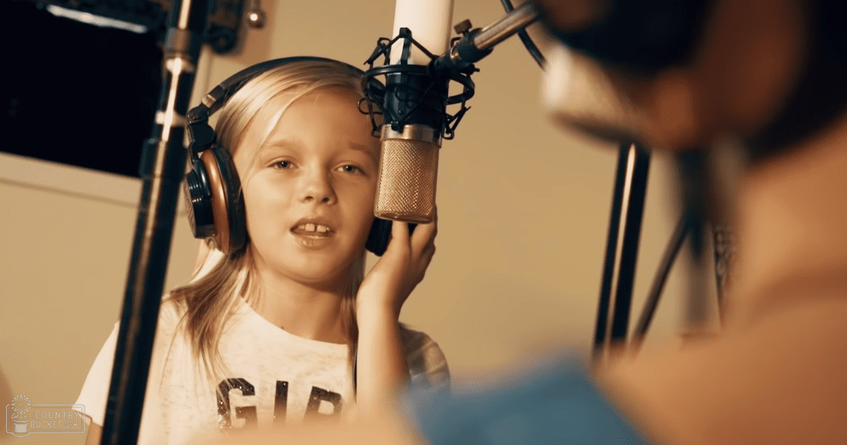 A young female in a recording studio wearing a 'GIRL' t-shirt, surrounded by audio equipment and another person assisting her.