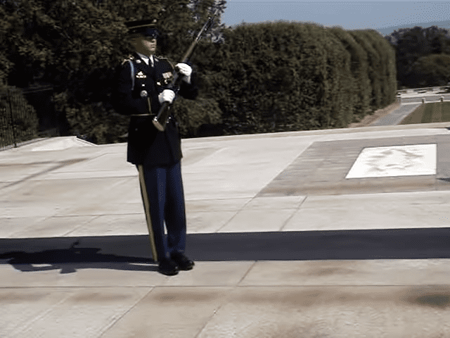 A soldier in a military uniform stands at attention before an eternal flame at Arlington National Cemetery on a sunny day with green surroundings.