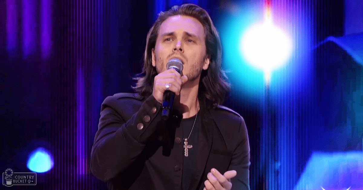 An adult male performer wearing a dark blazer and black shirt, holding a microphone on an indoor stage illuminated by dramatic blue and white lights.