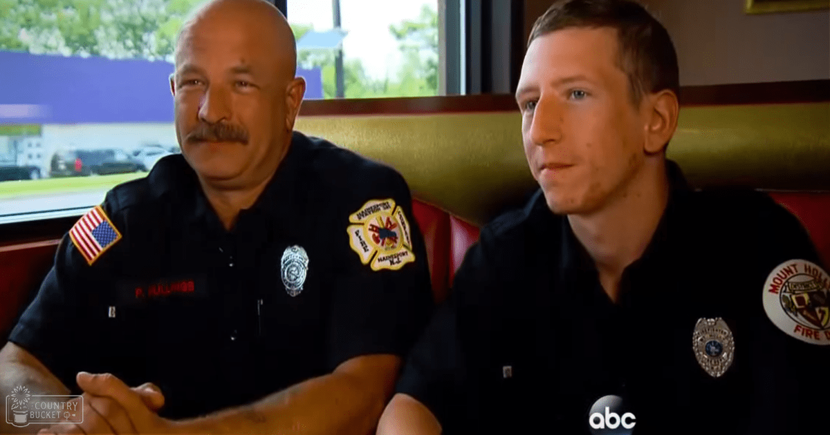 Two middle-aged male firefighters in dark blue uniforms discussing at red cushioned seating area within a restaurant-like setting with natural daylight.