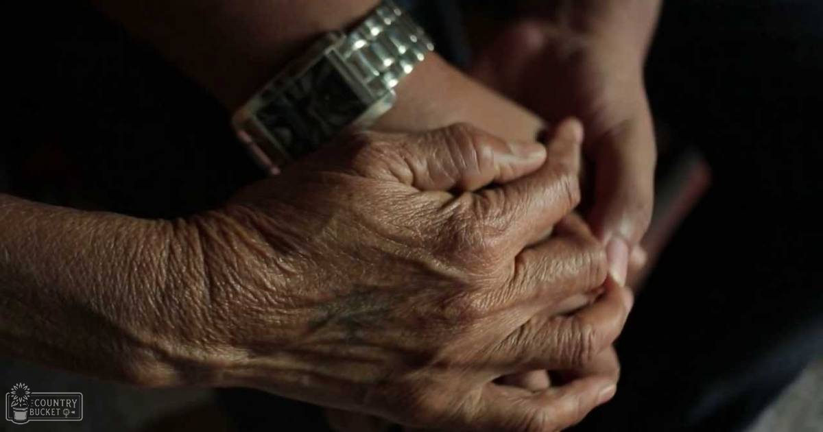 Close-up interaction between two individuals, likely indoors; older adult with wrinkled hands, younger adult wearing a watch on their wrist in dimly lit environment.