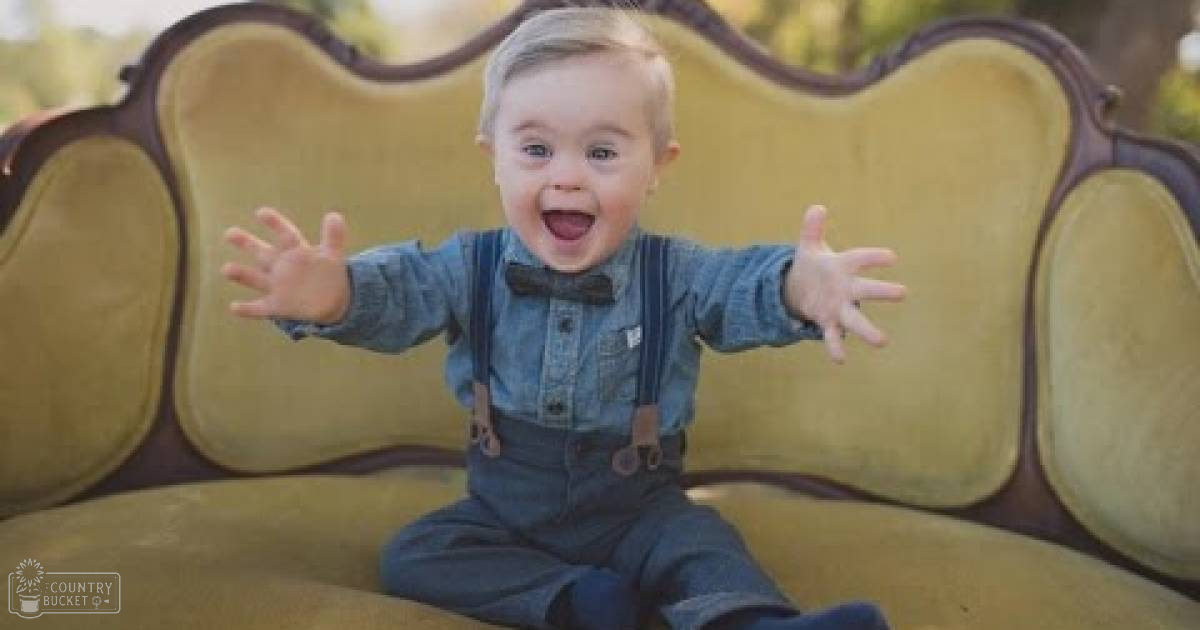 A toddler wearing a denim shirt with suspenders and a bow tie, outdoors in a park-like setting with a vintage yellow couch.