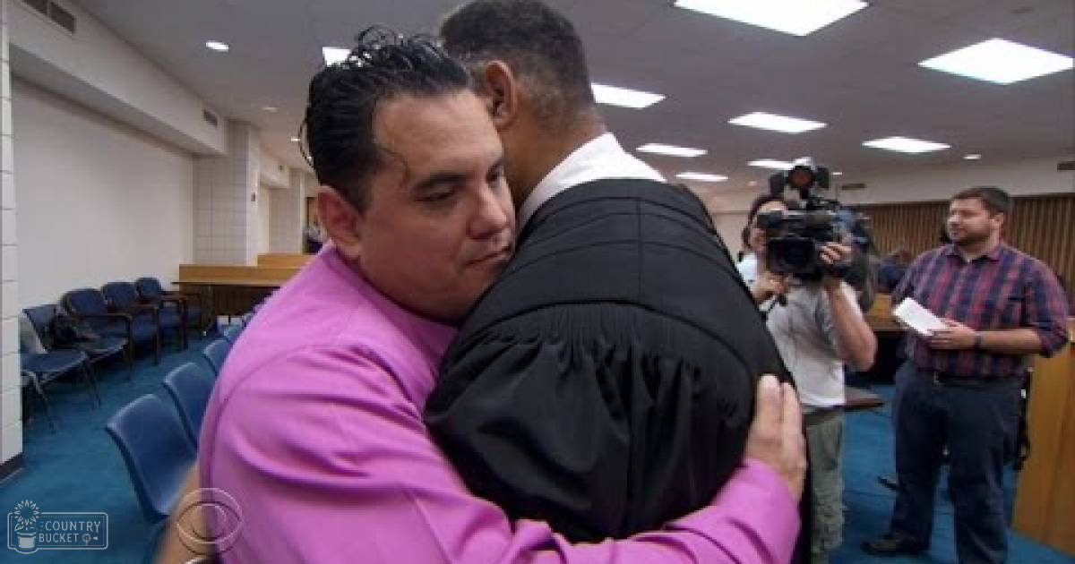 Adult male in a bright pink long-sleeved shirt hugging another individual wearing a black judicial robe in an indoor courtroom setting with fluorescent lighting.