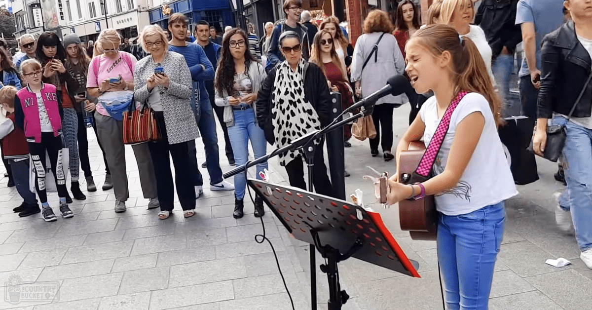 A young girl, child aged 8-10, performs with a brown acoustic guitar in an urban public space.