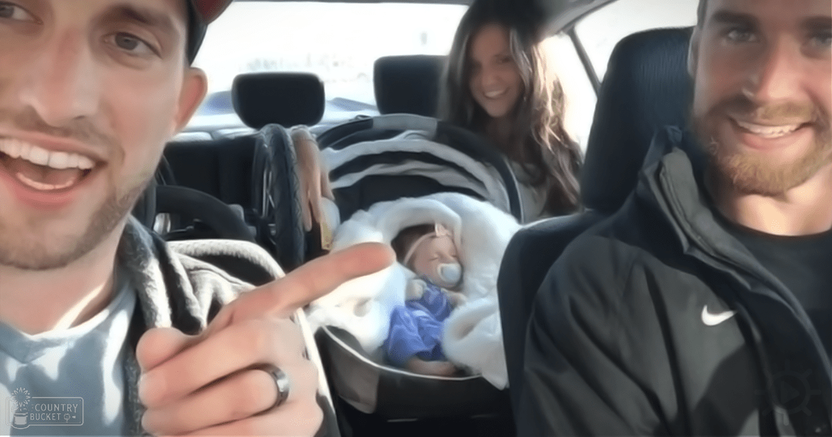 An infant wrapped in white blankets and wearing a blue outfit is seated in a car seat with two adults sitting inside, one wearing a dark jacket over a light-colored shirt or hoodie, the other in a black Nike jacket with facial hair. The scene takes place inside a vehicle during daytime.