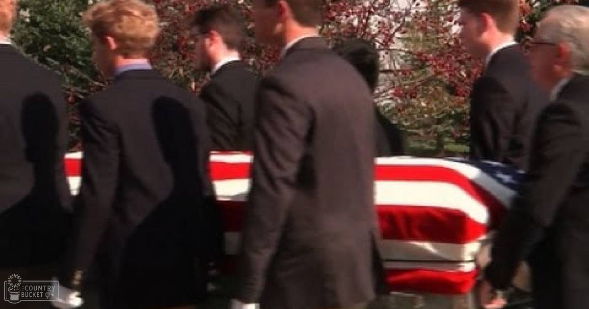 An outdoor autumn funeral procession with American flags draped over a casket, with middle-aged adults in dark suits against a backdrop of red-leaved trees and green foliage.
