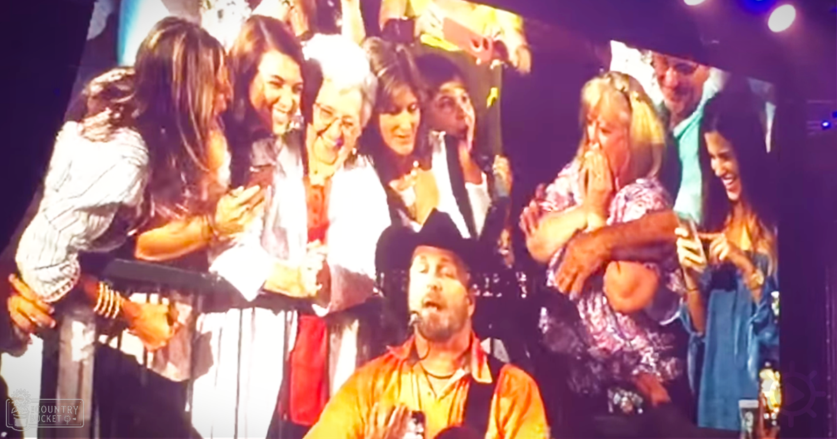 A musical performance with a bearded man in a red shirt and suspenders holding a microphone, surrounded by several women interacting warmly in an indoor venue under dim stage lighting.
