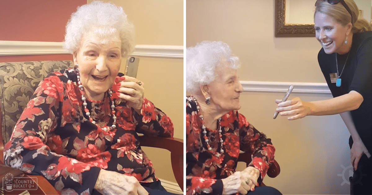 An older adult woman in a floral blouse with red flowers, sitting on an ornate chair indoors at a nursing home. A younger adult staff member is present nearby.