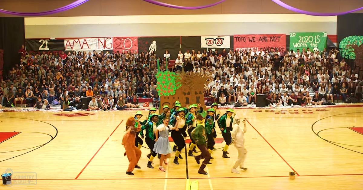 A school assembly in a gymnasium with performers dressed as characters from 'The Wizard of Oz,' including Dorothy and the Scarecrow, surrounded by an audience seated on bleachers.