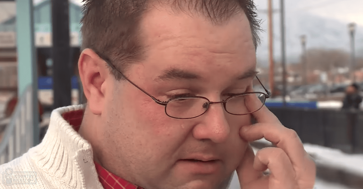 Middle-aged man wearing a white, collarless sweater with long sleeves and thin-framed glasses, holding something small to his ear against an urban backdrop during winter.