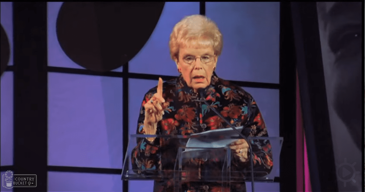 An older adult woman giving a speech at a podium in what seems to be a conference or event setting, wearing a loose-fitting floral blouse with long sleeves and glasses.