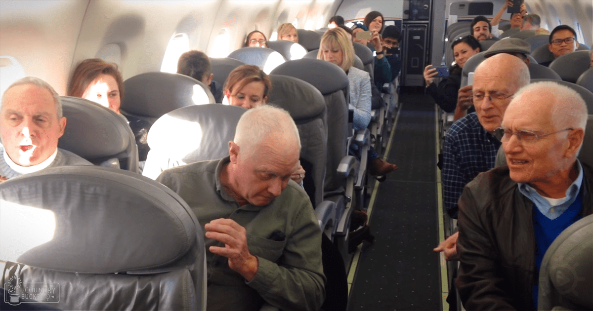 A group of older adults are seated and interacting in an airplane cabin interior during daytime flight.