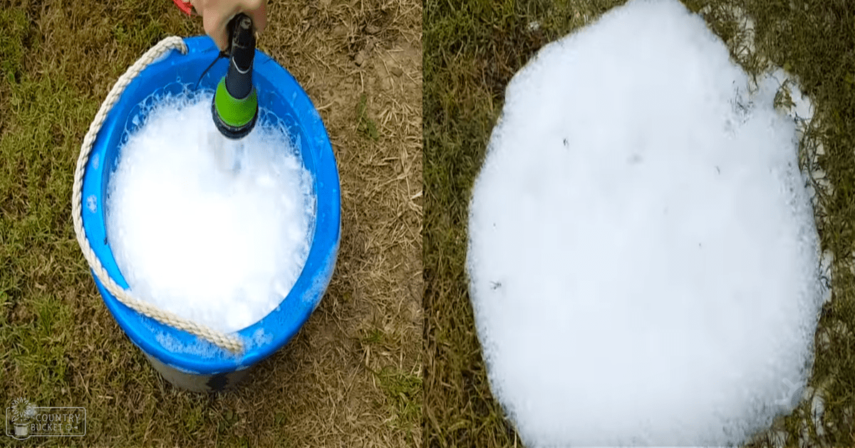 A bucket with soap suds being filled from a green and black hose on grass, likely an outdoor spring or summer scene.