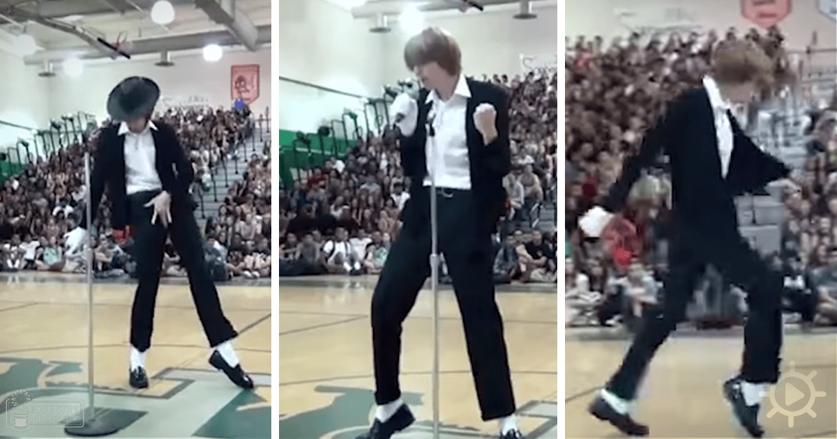 A young adult male in a black suit jacket and white shirt dances in an indoor gymnasium filled with spectators.