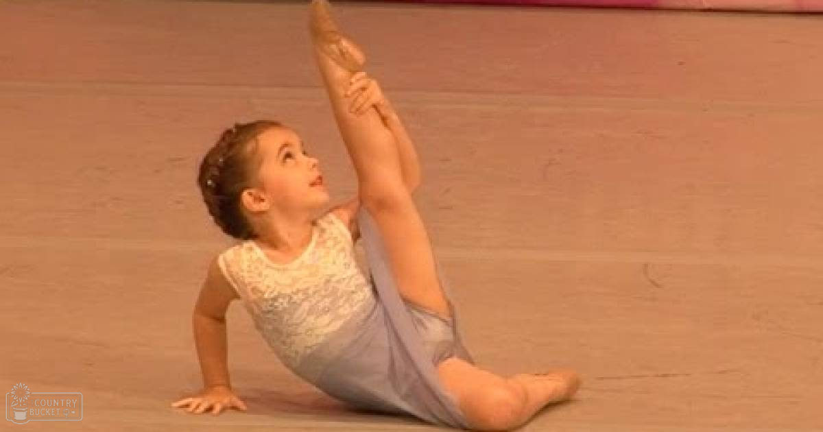 A toddler in a light-colored leotard practicing gymnastics indoors on a wooden floor, possibly during summer or spring.