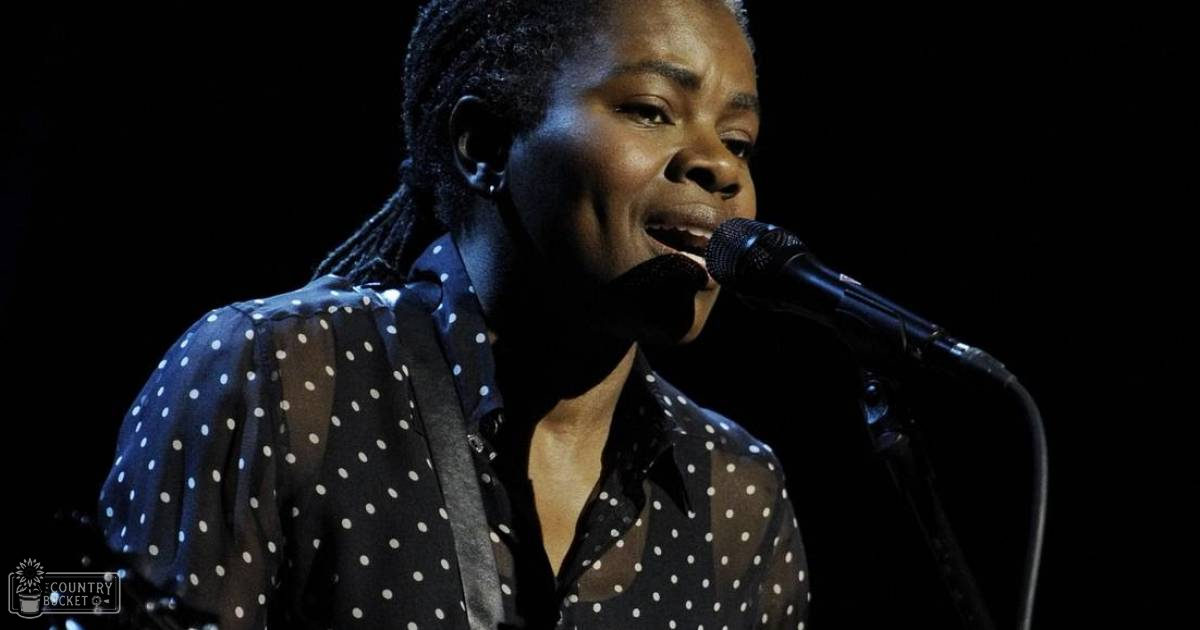 An adult female performer singing into a microphone at an indoor event with a polka-dotted blouse in black and white, contemporary style.