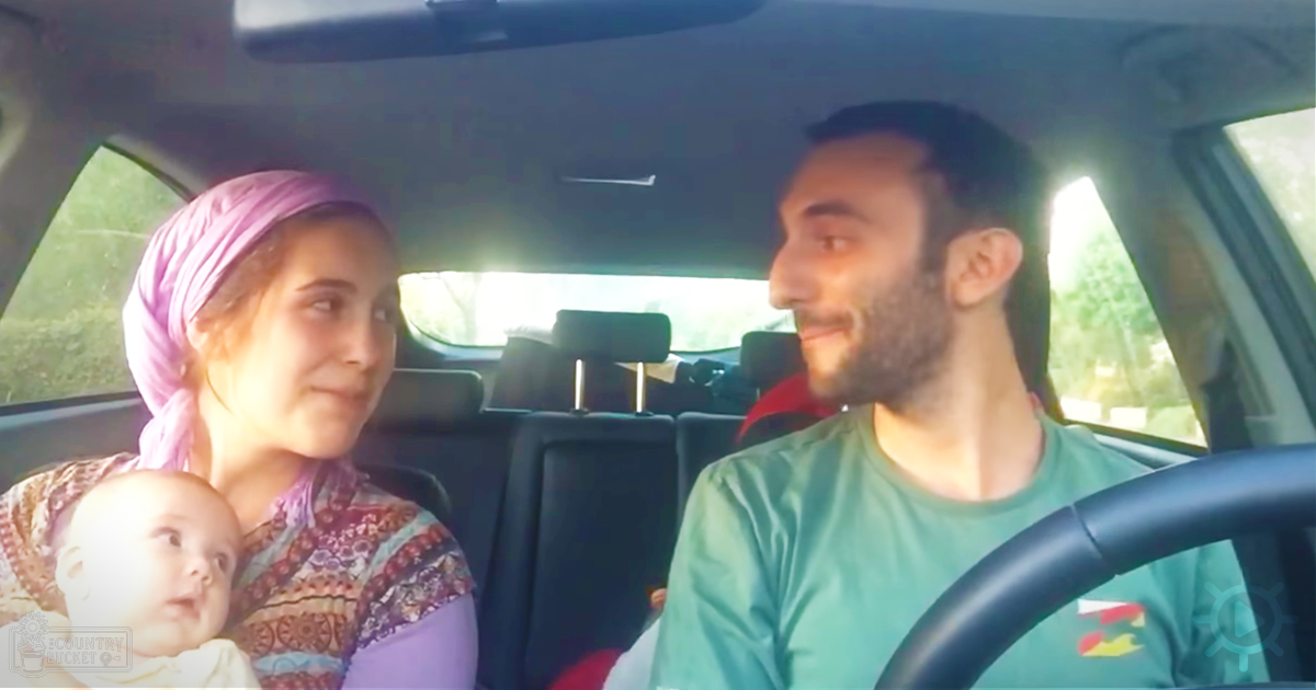 A family of three inside a car, with an adult female driver wearing a green t-shirt and an adult male passenger in a purple headscarf and patterned top. The infant looks curious while dressed in white.